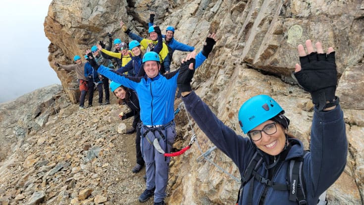 A group of people wearing helmets and harnesses stand on a rocky mountain trail, smiling and raising their arms toward the camera.